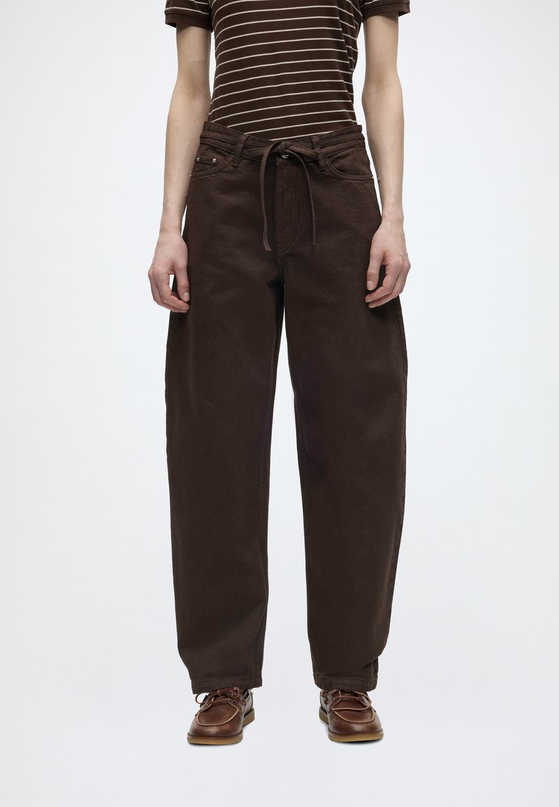 Άτομο που φοράει φαρδιά σκούρα καφέ παντελόνια με κορδόνι στη μέση, καφέ ριγέ κοντομάνικο πουκάμισο και καφέ δερμάτινα παπούτσια σε λευκό φόντο.