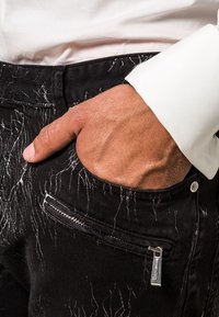 Jeans in denim neri con motivi striati bianchi, dotati di una tasca laterale con zip e una tasca frontale standard. Mano appoggiata sulla vita.