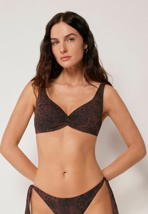 Frau mit langem dunklem Haar, die einen dunkelbraunen Bikini-Oberteil mit Leopardenmuster und passende Bikinihöschen mit seitlicher Bindung vor einem einfarbigen Hintergrund trägt.