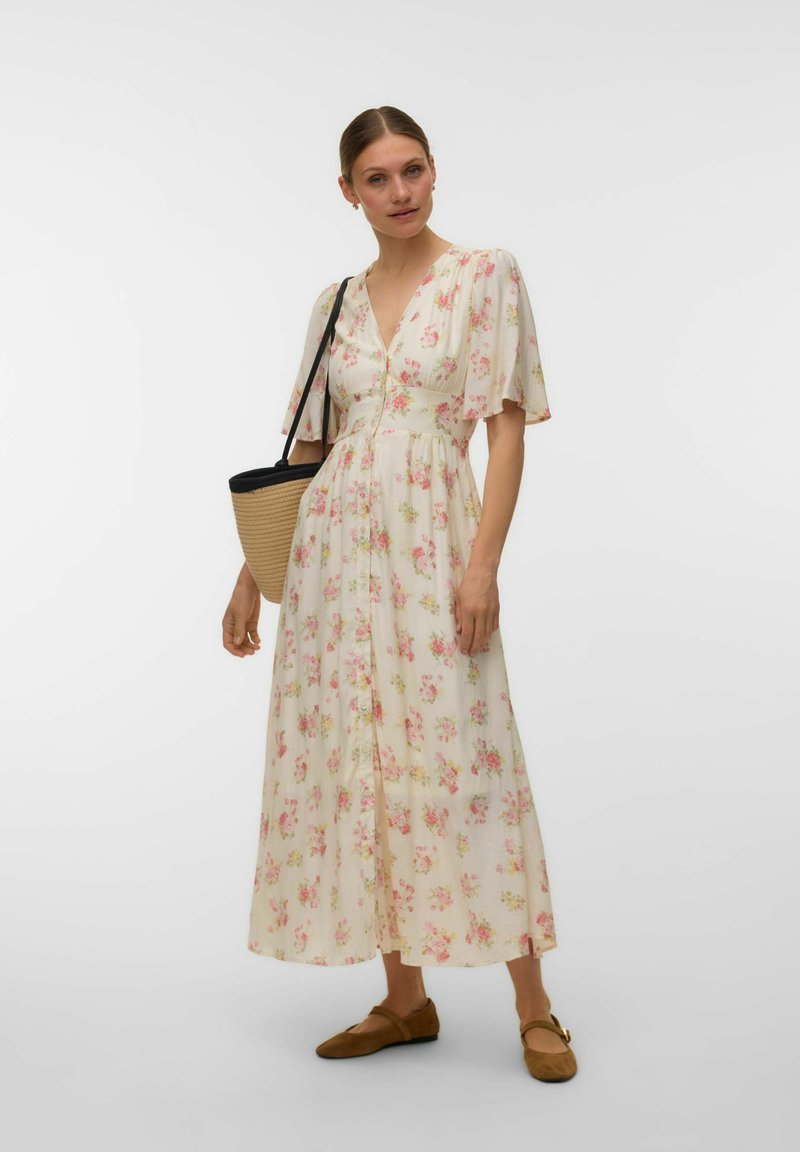 Femme portant une robe midi fleurie crème avec des manches volantées, des ballerines marron et un sac à bandoulière tressé, debout sur un fond blanc.