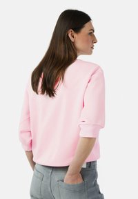 Rosa Sweatshirt mit 3/4-Ärmeln und rundem Ausschnitt, kombiniert mit grauen Hosen. Glatte Textur und minimales Design.