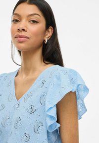 Gaiši zila īsām piedurknēm blūze ar V veida kakla izgriezumu, volāniem pie piedurknēm un smalkiem izšuvumiem ar izgriezumiem visā audumā.