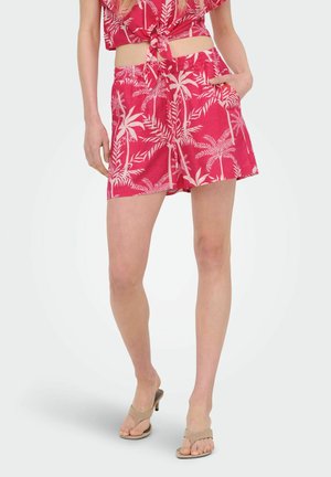 Vrouw draagt rode shorts met tropische palmprint en bijpassende gebonden crop top, met beige hakken sandalen, staande tegen een witte achtergrond.