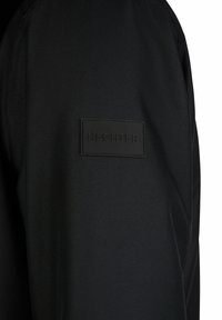 Mustmust kangasjakk, millel on tekstureeritud pind ja vasakul küljel on ristkülikukuju, reljeefne HECHTERi logo patch.