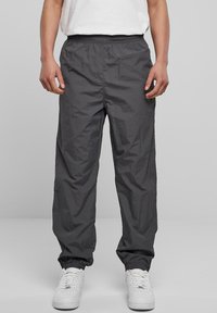 Person, der dunkelgraue Pants mit elastischem Bund und Knöchelbund sowie weiße Sneakers trägt, steht vor einem einfarbigen hellgrauen Hintergrund.