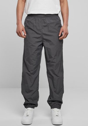 Persoon in donkergrijze elastische taille- en enkelbroek met witte sneakers, staand tegen een effen lichtgrijze achtergrond.
