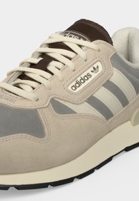 Κοντινό πλάνο ενός μπεζ και γκρι sneaker Adidas με λευκά κορδόνια, καφέ γλώσσα και το λογότυπο Adidas στην πλαϊνή πλευρά και τη γλώσσα.