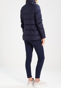 Donna che indossa una giacca a vento blu navy, pantaloni slim blu navy e sneakers bianche, in piedi con le spalle alla macchina fotografica su uno sfondo bianco.