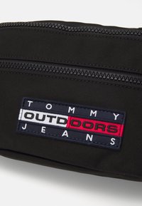 Svart stoffpose med glidelås og rektangulært merke. Merket viser "TOMMY OUTDOORS JEANS" i sterke farger: rød, hvit og marineblå.