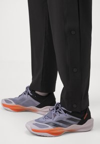 Person wearing black pants with side snap buttons and gray Adidas Adizero sneakers featuring orange accents.