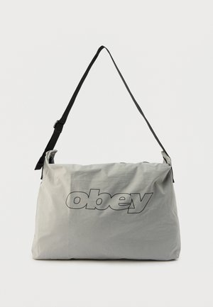 Lichtgrijze schoudertas met verstelbare zwarte riem, met een groot omlijnd "obey"-logo centraal aan de voorkant.
