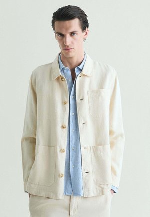 Joven con el cabello peinado hacia atrás lleva una chaqueta crema abotonada sobre una camisa azul claro, de pie contra un fondo claro y liso.