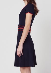 Marinblå stickad klänning med korta ärmar, röd vågig kant vid halsringning och midja, samt en plisserad kjoldesign.