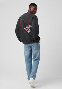 Sudadera gris oscura con un gran diseño de corazón rojo y alfileres de seguridad en la parte posterior, combinada con jeans de mezclilla azul claro y zapatillas blancas.