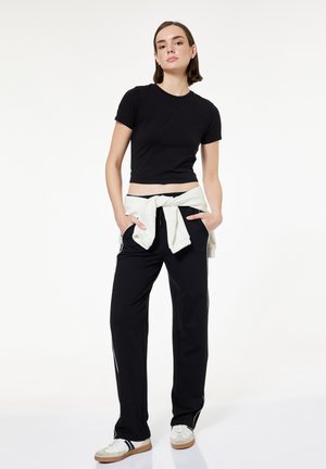 Vrouw staand met handen in de zakken, draagt een zwarte cropped top, zwarte trainingsbroek met witte strepen, wit sweatshirt gestrikt rond de taille en witte sneakers.