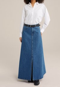 Džínová maxi sukně s předním rozparkem, modré barvy a vysokým pasem. V kombinaci s bílou košilí na knoflíky a černým opaskem pro kontrast.