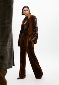 Blazer in velluto marrone con strisce a contrasto, abbinato a pantaloni larghi coordinati. Il modello indossa una camicia nera con un design grafico.