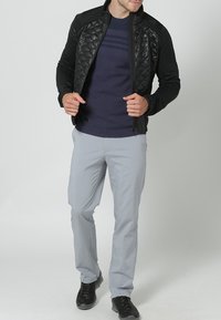 Black quilted jacket with a fitted design, worn over a navy blue sweater and light grey trousers. Black footwear completes the look.