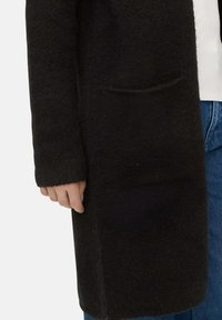 Schwarzer, strukturierter Wollmantel mit langen Ärmeln; verfügt über eine kleine, vordere Tasche und einen geraden Schnitt, kombiniert mit blauen Jeans.