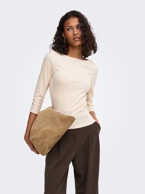 Vrouw met krullend donker haar die een beige top met lange mouwen en donkerbruine broek draagt, en een grote lichtbruine suède clutch vasthoudt.
