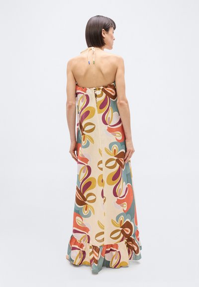 Γυναίκα με κοντά καστανά μαλλιά, φορούν μια πολύχρωμη μακριά φόρεμα με λουλούδια χωρίς τιράντες και βολάν στο τελείωμα, στέκεται γυρισμένη ανάποδα μπροστά σε απλό φόντο.