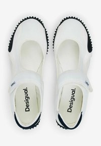 Des chaussures de style Mary Jane blanches avec des semelles en caoutchouc noires, dotées d'une sangle Velcro et de détails texturés sur la semelle et les côtés. Logo de la marque visible à l'intérieur.