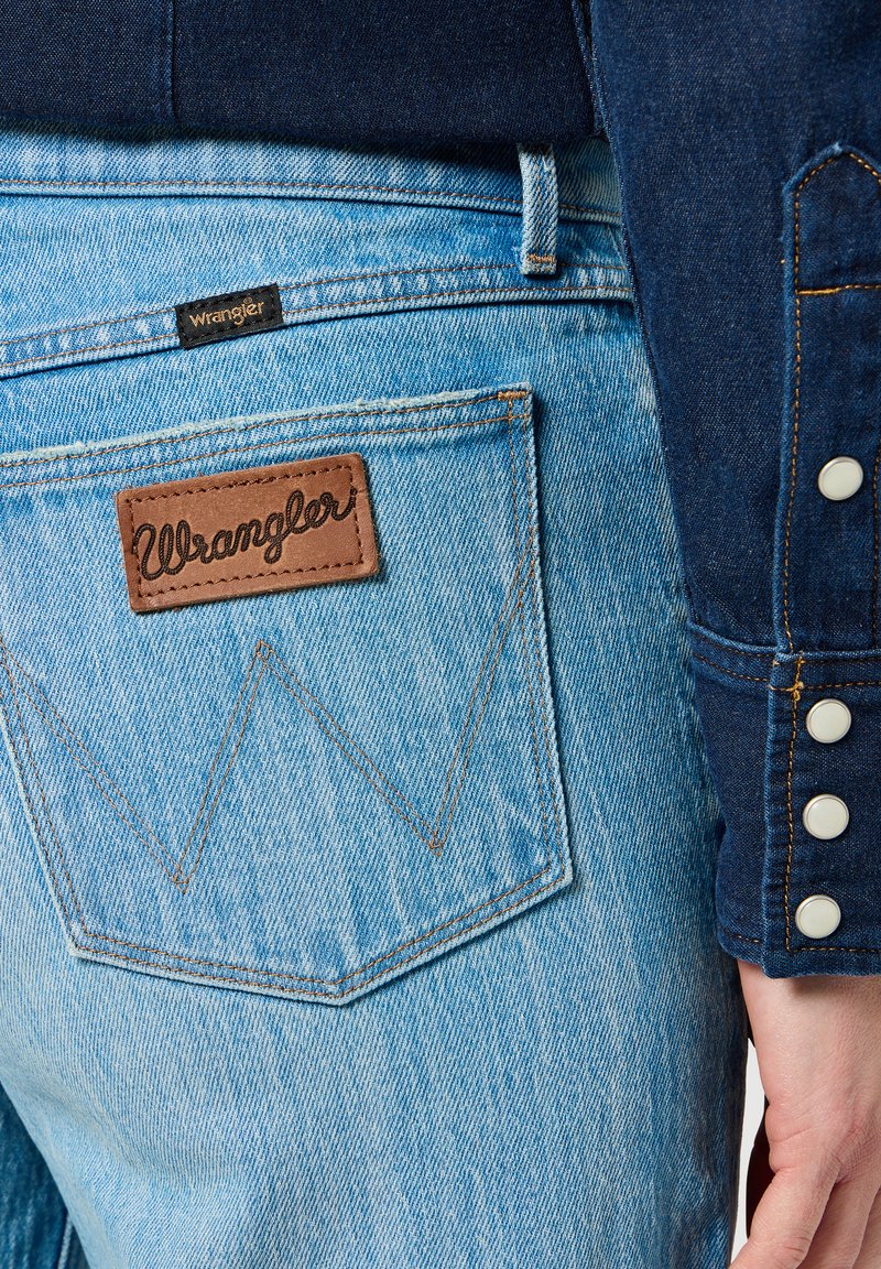 Jasnoniebieskie dżinsy z brązową skórzaną łatą z napisem "Wrangler" na tylnej kieszeni oraz kontrastowym przeszyciem. Obok ciemnoniebieska koszula dżinsowa z zatrzaskami.