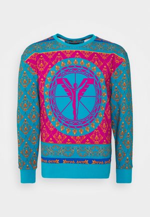 Sweat-shirt à manches longues turquoise et magenta avec des motifs dorés ornés et un motif circulaire central présentant une silhouette humaine stylisée avec des ailes.