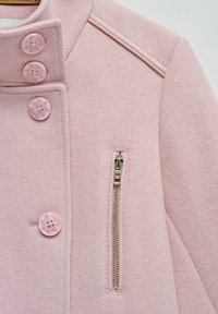 Chaqueta de patrón de espiga en color rosa con cuatro botones grandes de color rosa, un bolsillo con cremallera lateral y un diseño de cuello estructurado.