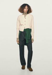 Cardigan rosa chiaro con bottoni in legno, maglione verde legato in vita, scarpe in suede marrone e pantaloni in denim scuro. Design semplice e moderno.