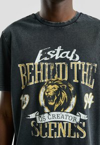 Czarna bawełniana koszulka z przetartym białym i złotym nadrukiem graficznym, zawierająca ilustrację lwa i tekst "Estab Behind The Scenes".
