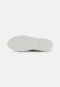 Suola della scarpa in materiale gommoso bianco testurizzato con un motivo ondulato e un piccolo accento di branding vicino al centro.