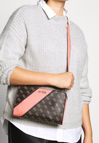 Sort og brunt mønstret håndtaske med koralfarvet diagonalt accent, med lynlås og metal hardware, holdt op mod en grå sweater.