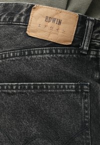 Črne denim hlače z teksturirano površino, opremljene s svetlo rjavim usnjenim oznako z embossed napisom "EDWIN" ter šivi na pasu in žepih.