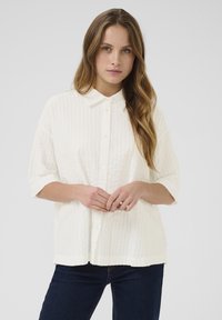 White, textured button-up shirt with a collar, short sleeves, and a relaxed fit, paired with dark blue jeans.