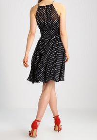 Schwarzes gepunktetes Kleid mit einem Halterneck und einem schwingenden Rock. Durchsichtiger Oberteil mit strukturierten Akzenten und roten High Heels.