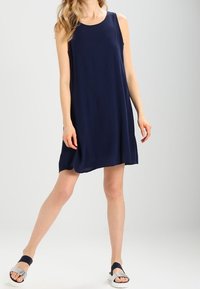 Mekko, jossa ei ole hihoja, merensinisellä värillä, polven mittainen, pehmeä kangas, A-linjainen muoto. Yhdistetty mustilla hihnoilla varustettuihin sandaaleihin, joissa on metallinhohtoisia hopeasävyjä.