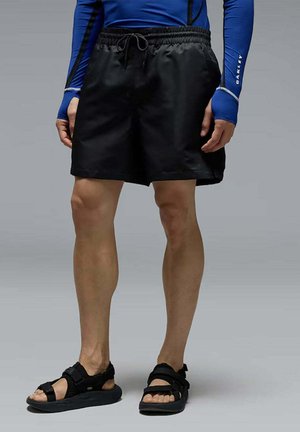 Persona che indossa pantaloncini neri con coulisse, sandali neri e una maglia blu a maniche lunghe con fori per il pollice, in piedi su uno sfondo grigio.
