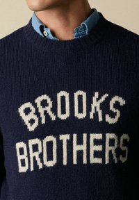 Maglione a maglia blu navy con testo bianco "BROOKS BROTHERS". Collo rotondo con dettagli a costine. Indossato sopra una camicia collare azzurro chiaro.