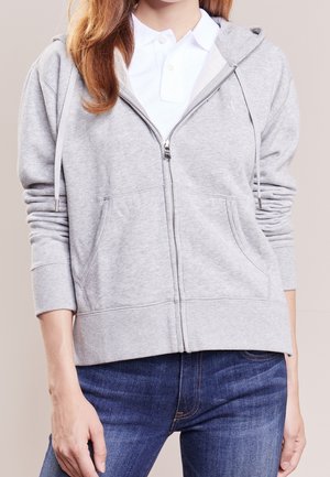 Jeune femme portant un sweat à capuche zippé gris clair par-dessus un polo blanc et un jean bleu, debout sur un fond neutre.