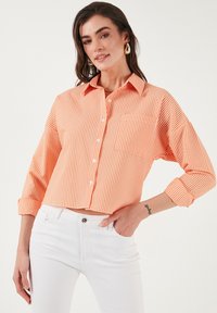 Oranje en witte gestreepte cropped shirt met een knoopsluiting aan de voorkant, kraag en opgerolde mouwen. Draagt bij witte jeans met een voorzak.
