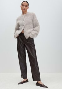 Woman with slicked-back hair wears a fuzzy beige cardigan, high-waist dark brown leather pants, and matching pointed flats, standing on a white floor.