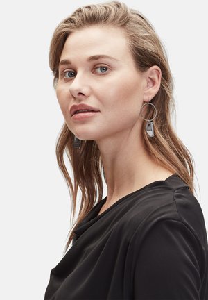 Des boucles d'oreilles en argent en forme de créoles présentent des accents rectangulaires ; le modèle porte un haut noir avec un décolleté drapé et a de longs cheveux ondulés séparés sur le côté.