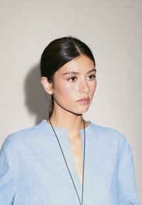 Blouse en lin bleu clair avec un décolleté en V et des manches longues, tissu texturé, et un collier fin en cordon noir. Fond neutre.