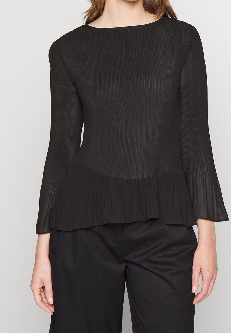 Femme portant un chemisier plissé noir à manches longues avec un ourlet légèrement évasé, associé à un pantalon noir ample, debout devant un fond uni.