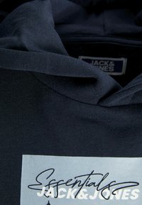 Haut à capuche bleu marine en coton, avec un patch logo rectangulaire bleu clair affichant le texte noir "Essentials JACK&JONES." Texture douce.
