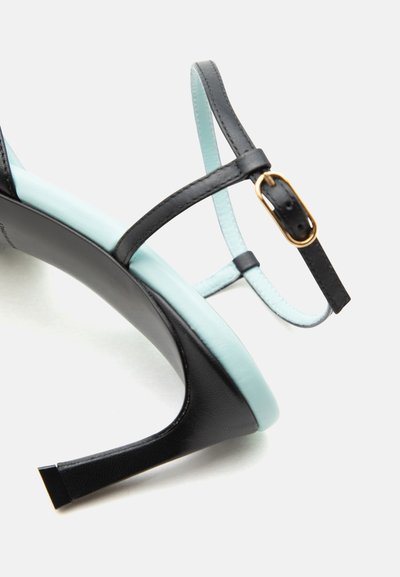 Μαύρο πέδιλο με ψηλό τακούνι και λεπτές λουρίδες, χρυσή αγκράφα και επενδεδυμένη εσωτερική σόλα γαλάζιου χρώματος, σε λευκό φόντο.