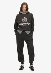 Dark grey sweatshirt with printed "Superdry" logo, layered over a blue and white striped shirt, paired with matching sweatpants and loafers.
