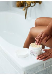 Weißer runder Behälter mit der Aufschrift "perfekte Beine 100% natürlicher Peeling" und strukturiertem Deckel, auf der geschwungenen Kante einer weißen Badewanne platziert.