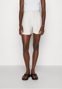 Pantalones cortos color crema con rayas finas y cintura elástica, confeccionados en tela ligera. Combinados con sandalias negras tipo flip-flop.